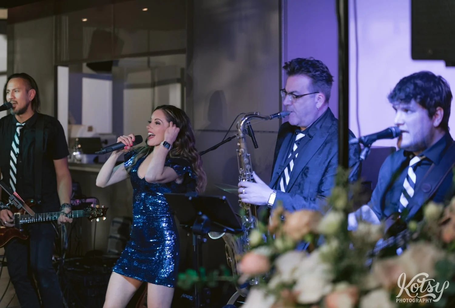 A woman in a blue sequin dress is singing into a microphone with a band featuring a guitar player saxophone player and bass guitarist during a Second Floor Events wedding reception in Toronto, Canada.