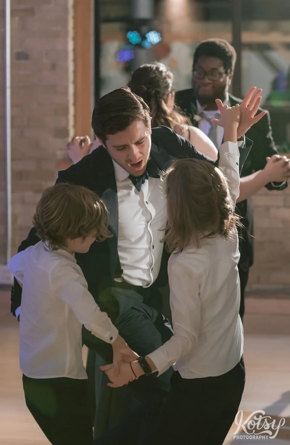 A man in a black suit and black bow tie dances with two children in white dress shirts during a Second Floor Events wedding reception in Toronto, Canada.