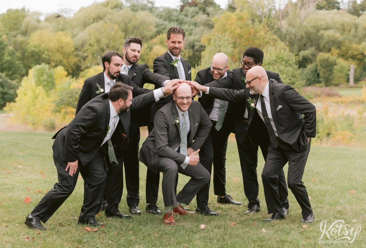 A groom wearing a green tie and gray suit squats as his seven groomsmen placed their hands on his head at West Deane Park in Toronto, Canada