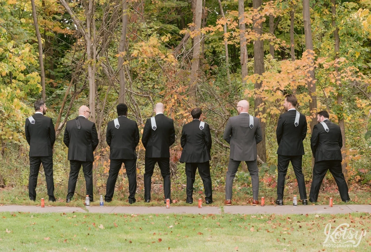 A groom and his 7 groomsmen stand in a line facing away from the camera and ties over their shoulder mimicking Urinating on the grass at West Deane Park in Toronto, Canada