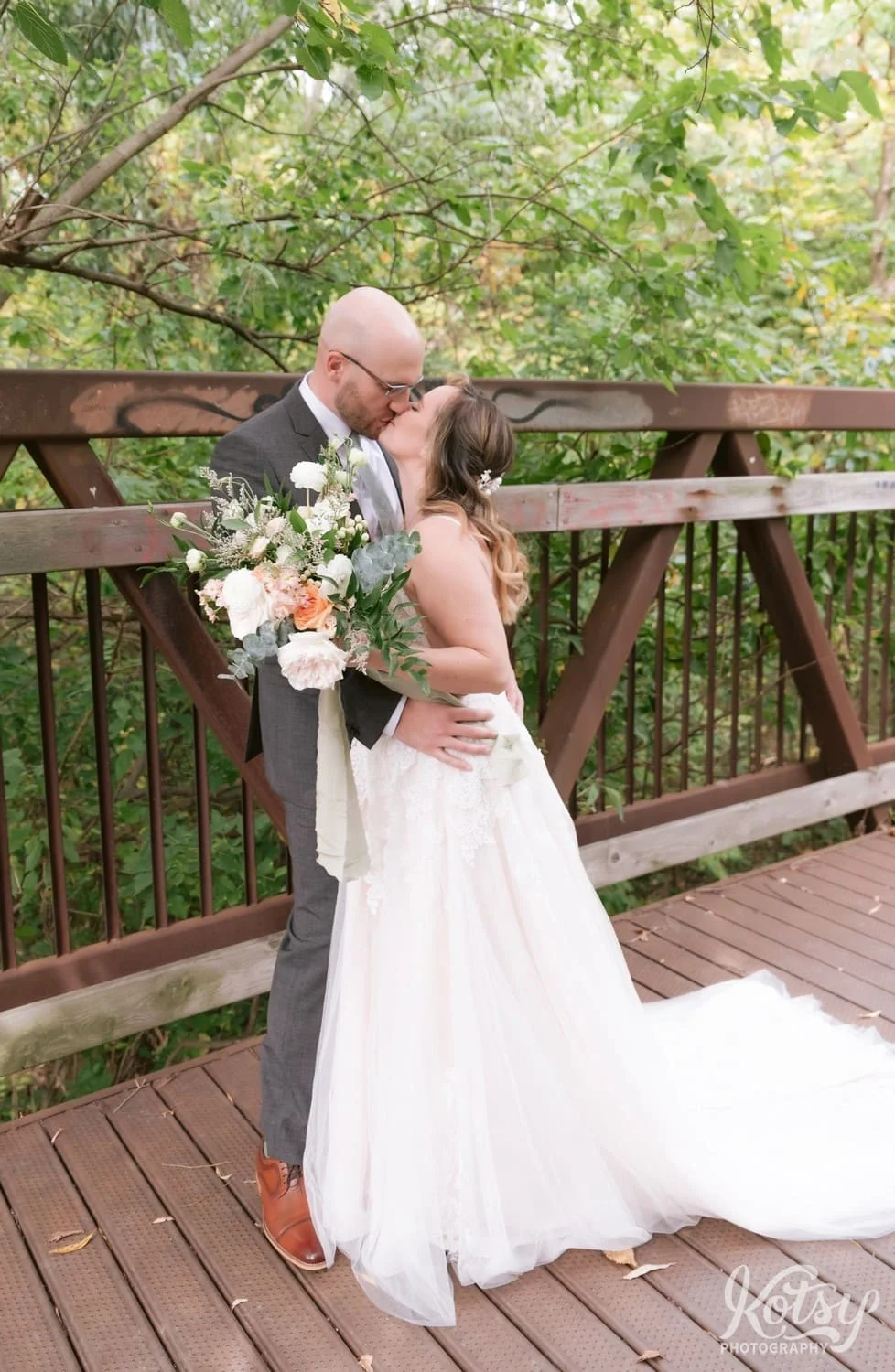 A bride wearing a white bridal gown and a groom wearing a gray suit kiss on a bridge during their first look at West Deane Park in Toronto, Canada