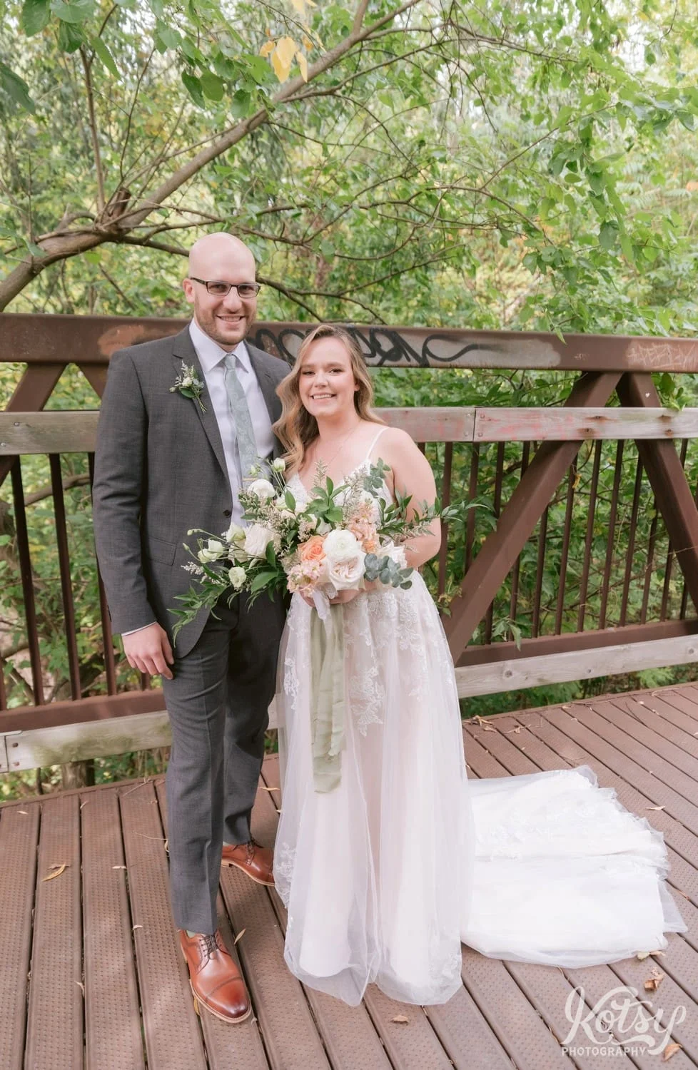 A bride wearing a white bridal gown, holding a flower bouquet and a groom wearing a Gray suit pose for a photo on a bridge during their first look at West Deane Park in Toronto, Canada