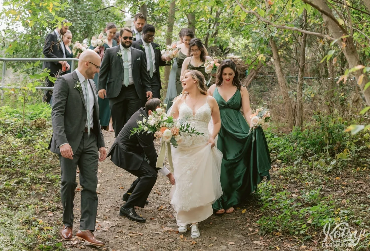 A bride and a white bridal Gown holding a flower bouquet lets out a big laugh as she walks down a dirt path with her groom and wedding party at West Deane Park in Toronto, Canada