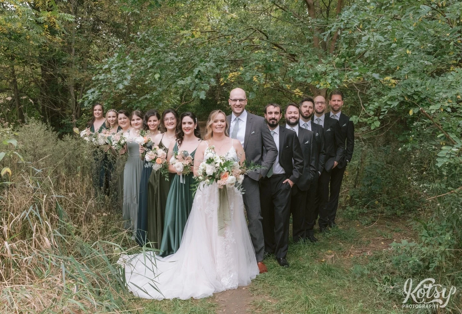 A bride groom and wedding party complete with flower bouquets white bridal gown and green bridesmaid dresses black groomsmen suits and gray suit on groom pose for a group photo and a triangular pattern at West Deane Park in Toronto, Canada