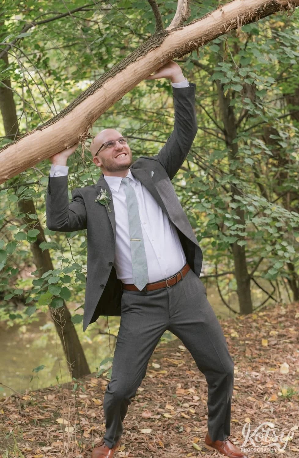 A groom wearing a white suit and green tie holds his hands up in the air pretending to push a large tree branch up at West Deane Park in Toronto, Canada