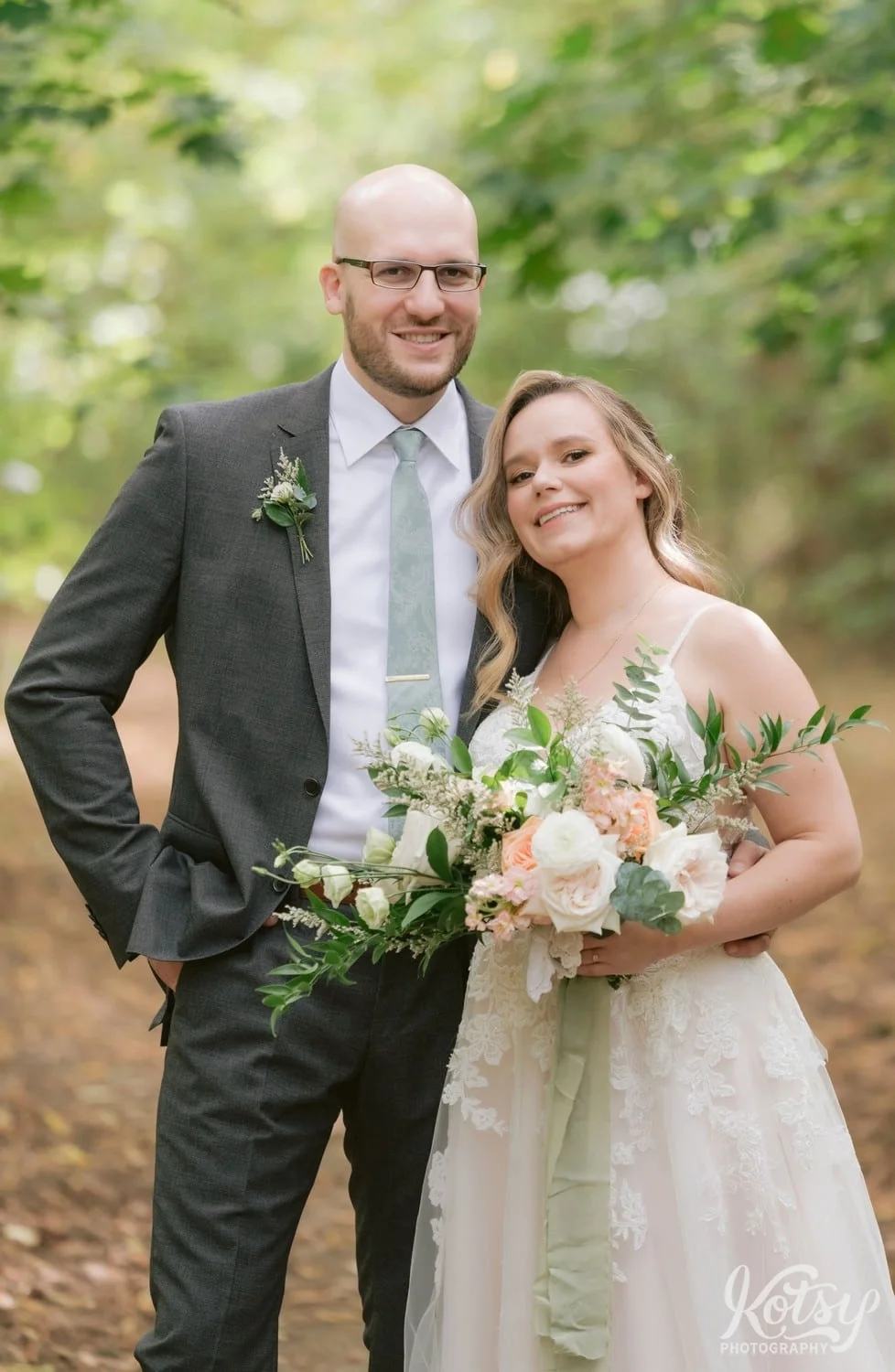 A grim wearing a gray suit and green tie and a bride wearing a white bridal gown and holding a flower bouquet pose for a photo at West Deane Park in Toronto, Canada