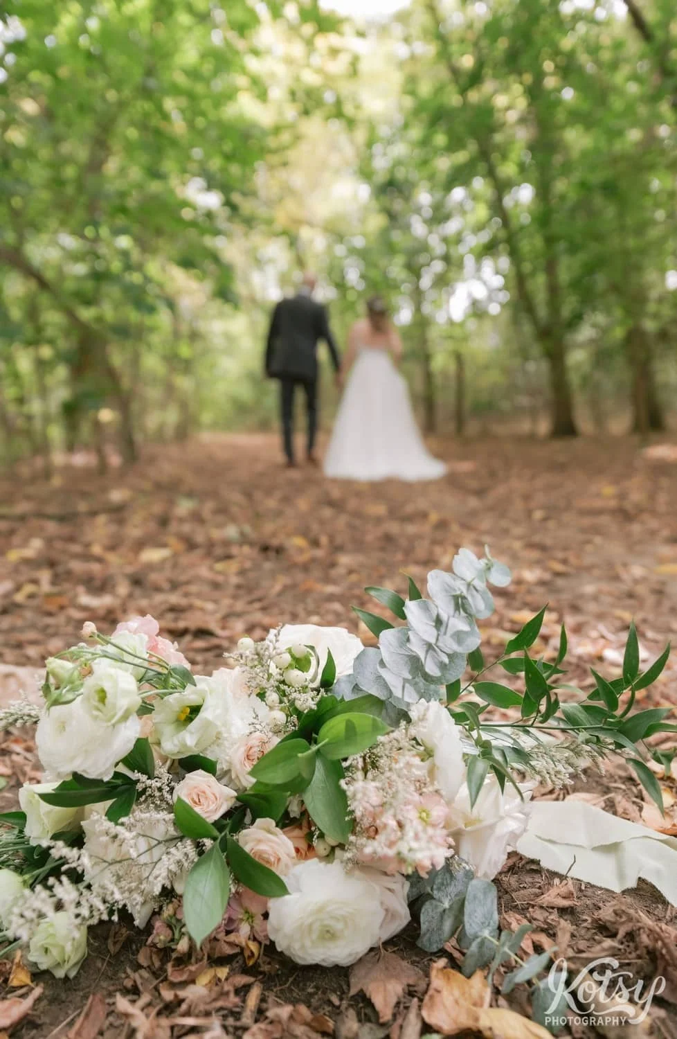 A close up shot of a flower bouquet on lead covered dirt path at West Deane Park in Toronto Canada. Seen in the background is a bride and groom walking away from the camera while holding hands