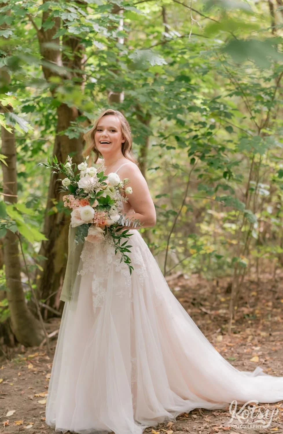 A bride holding a flower bouquet and wearing a white bridle of gown poses for a photo on a dirt leaf covered half at West Deane Park in Toronto, Canada