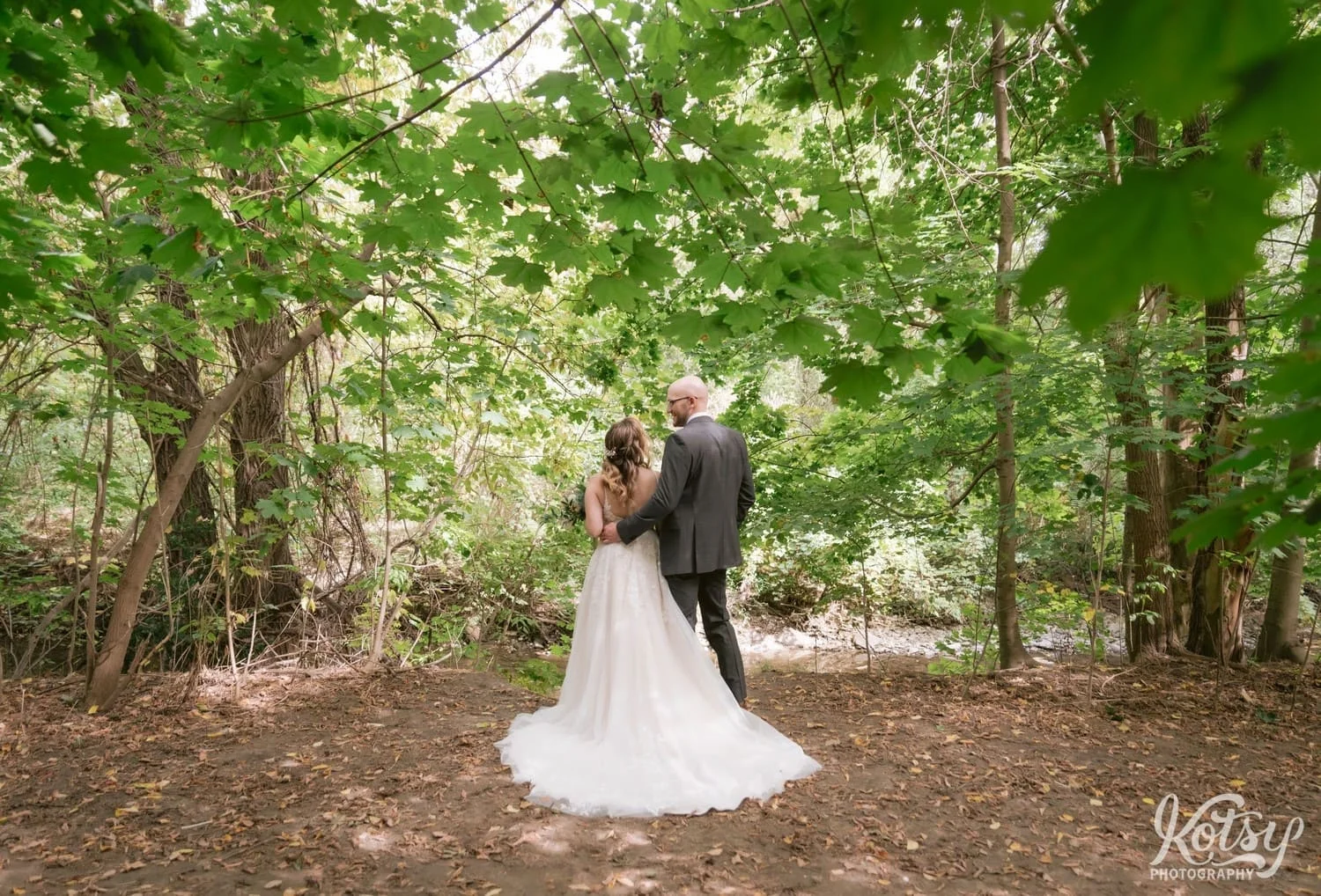 A wide shot of a groom in a gray suit holding his bride in a white bridal gown looking away from the camera at West Deane Park in Toronto, Canada