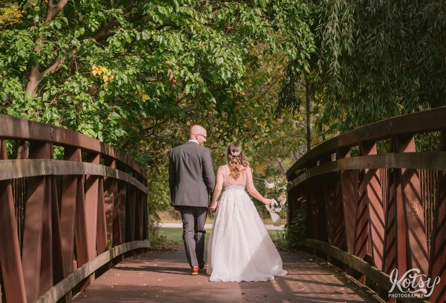 A real shot of a groom wearing a gray suit and a bright wearing a white vital gown holding a flower bouquet walking away from the camera on a steel bridge at West Deane Park in Toronto, Canada