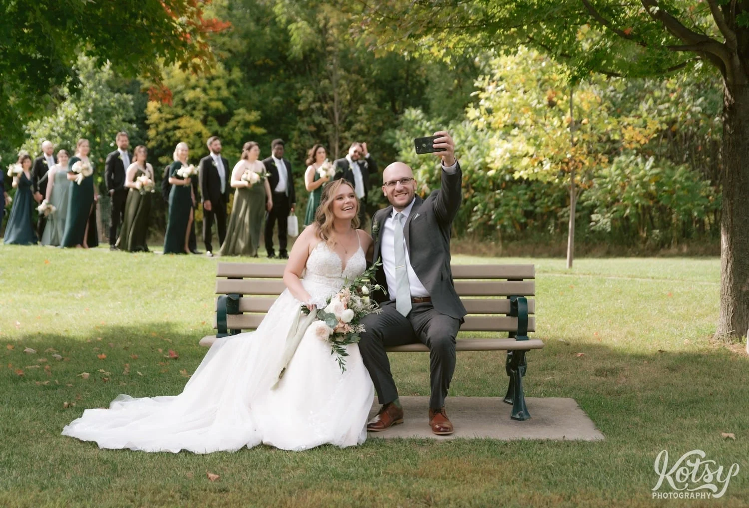 A groom wearing a gray suit and green tie holds a phone to take a selfie with his bride wearing a white bridal gown and holding a flower bouquet on a park bench while their wedding party walks behind them on a path at West Deane Park in Toronto, Canada