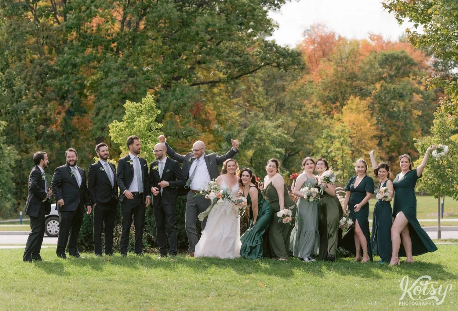 A groom wearing a gray suit and a bride wearing a white bridal gown holding flowers dance and joke around with their wedding party wearing green and black at West Deane Park in Toronto, Canada