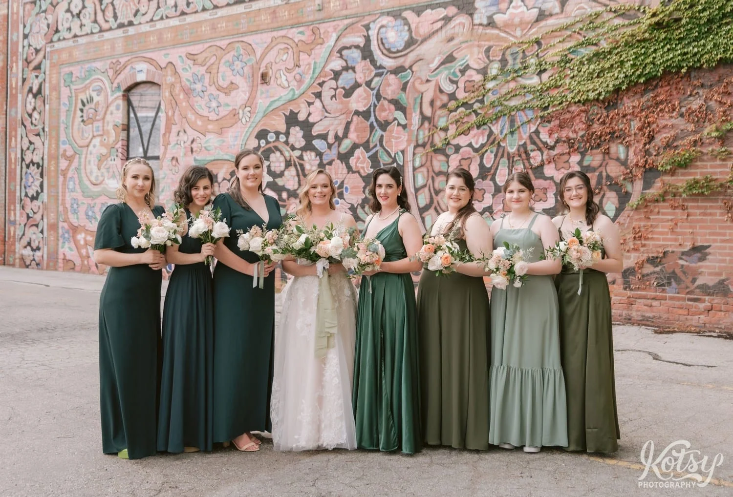 A bride wearing a white bridal gown poses with her bridesmaids holding bouquets of flowers in front of a mural outside the carpet factory in front of a colourful mural in Toronto, Canada