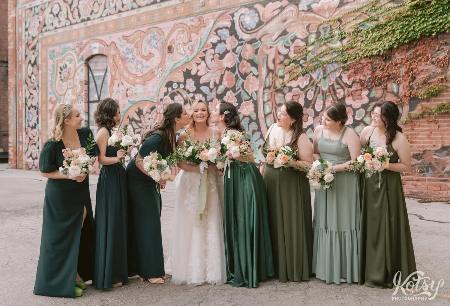 True bridesmaids holding flower bouquets mimic a kiss to a bride wearing a white bridal gown while the other bridesmaids look on outside the carpet factory in Toronto Canada in front of a colorful mural