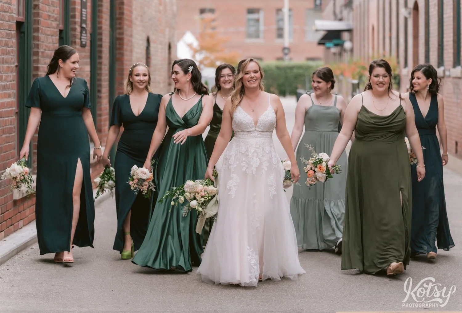 A bride wearing a white bridal gown and holding a flower bouquet walks with her bridesmaids wearing green dresses in between buildings at the carpet factory in Toronto Canada