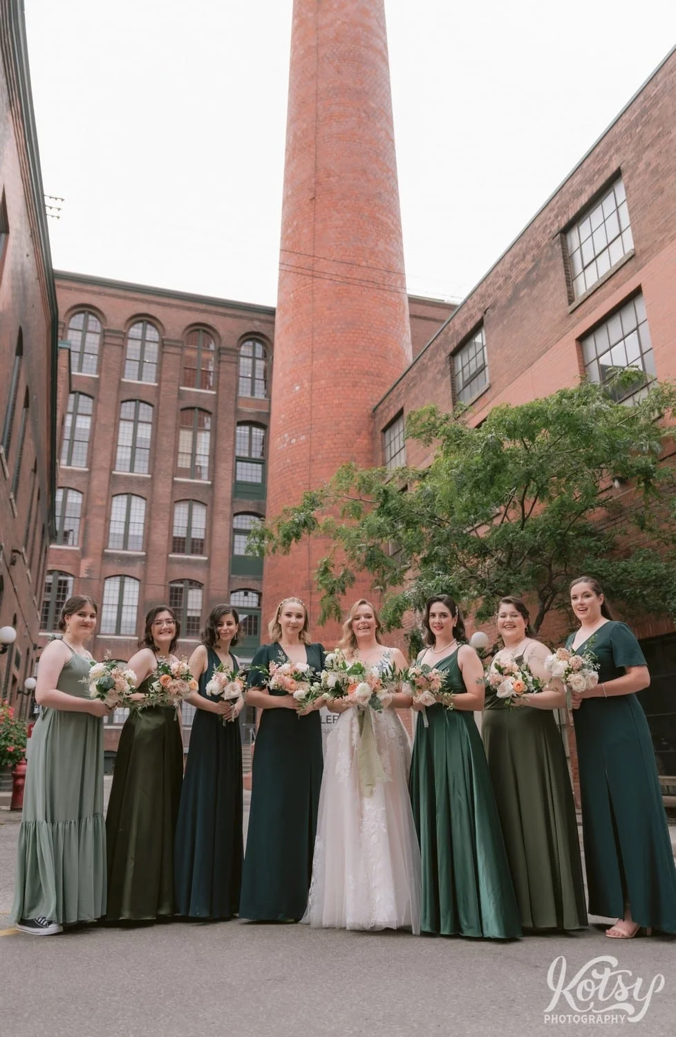 A bride wearing a white bridle gown holding flour bouquet poses for a group photo in front of a tall smoke stack with her bridesmaids wearing green dresses