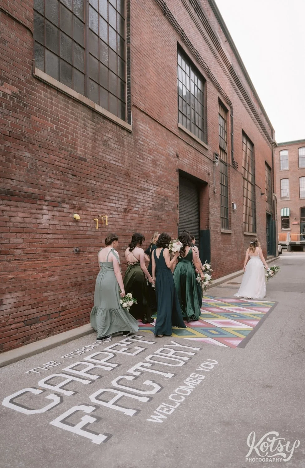 A shot of a bride wearing a white bridal gown holding a flower bouquet walks in front of her bridesmaids in green dresses over a carpet factory logo painted on the ground in Toronto Canada