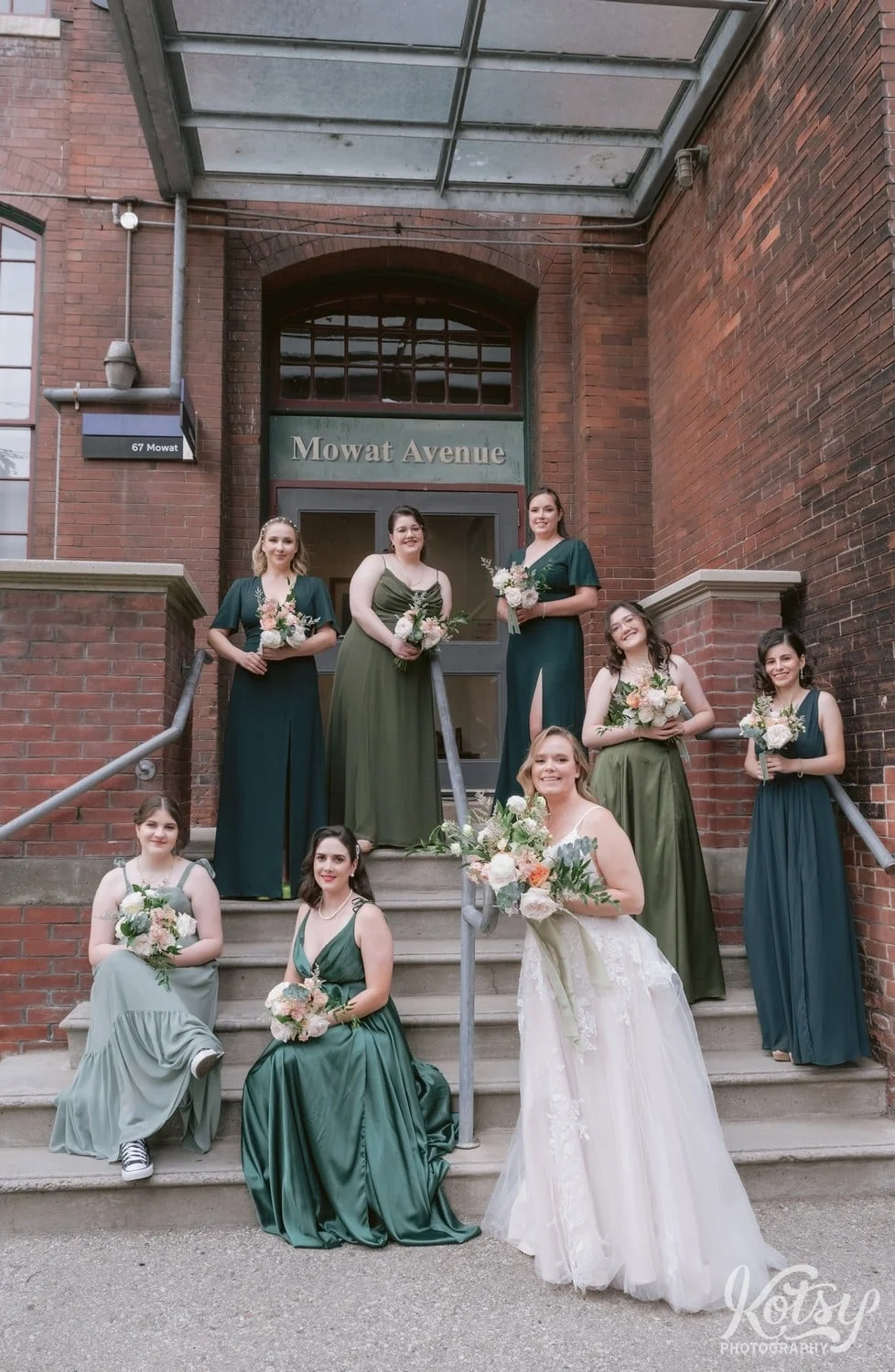 A bride wearing a white bridal gown and holding a bouquet of flowers poses with her bridesmaids wearing green dresses in front of a warehouse building at the carpet factory in Toronto Canada