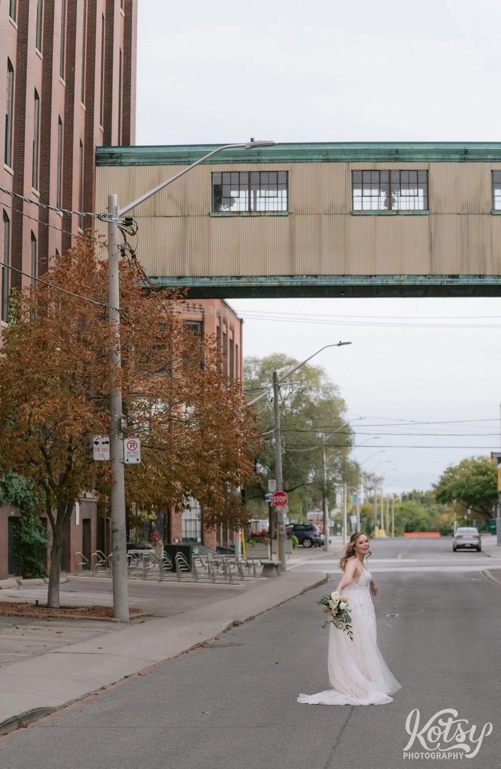 A bride wearing a white bridal gown and holding a bouquet of flowers looks back at the camera well laughing and standing in the middle of a empty road beside the carpet factory with pedestrian bridge in Toronto Canada