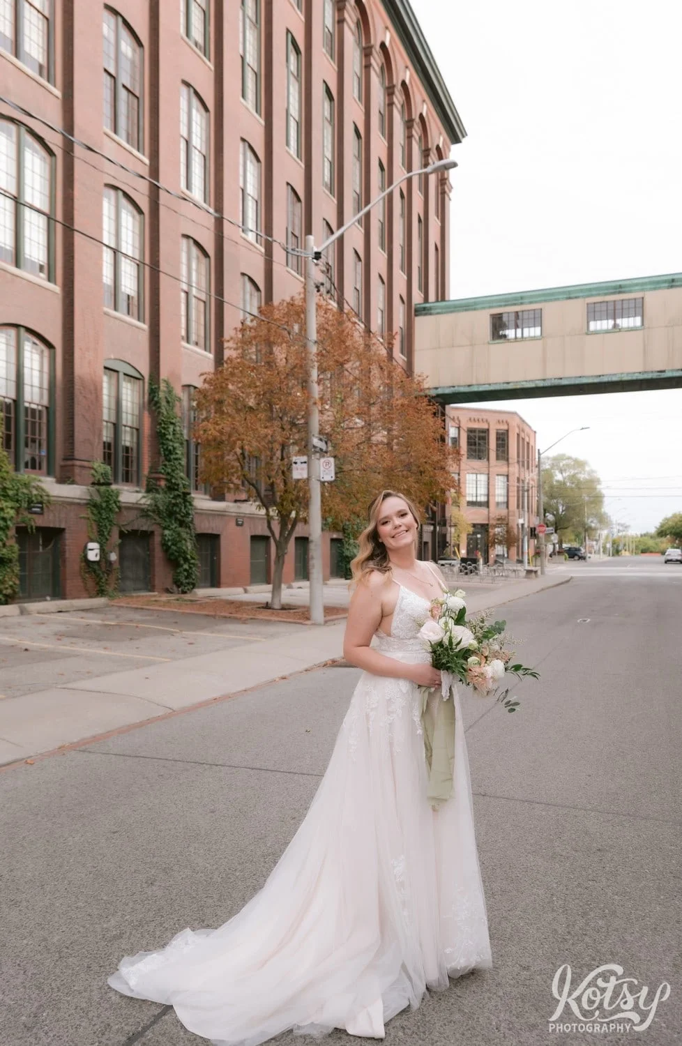 A bride wearing a white bridal gown and holding a bouquet of flowers poses in the middle of an empty road with the carpet factory and pedestrian bridge in the background in Toronto Canada