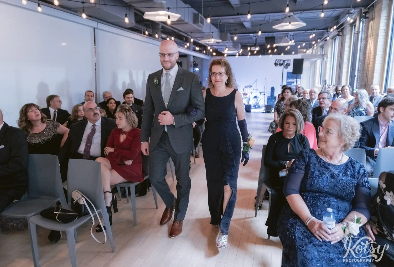 A groom wearing a gray suit and green tie walks down the aisle with his mother during his wedding ceremony at second floor events in Toronto Canada