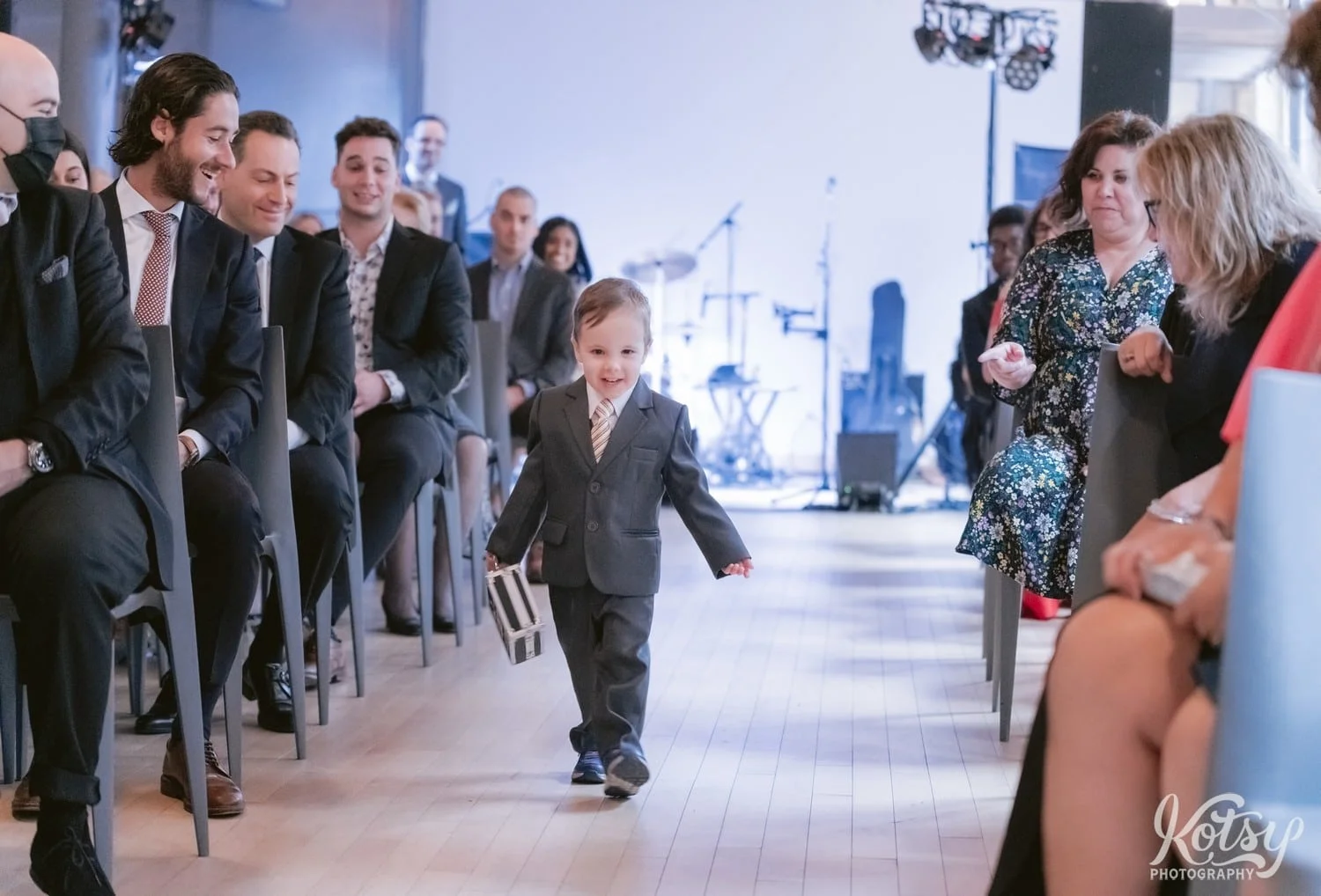 A ring bearer holding a little suitcase walks down the aisle during a wedding ceremony