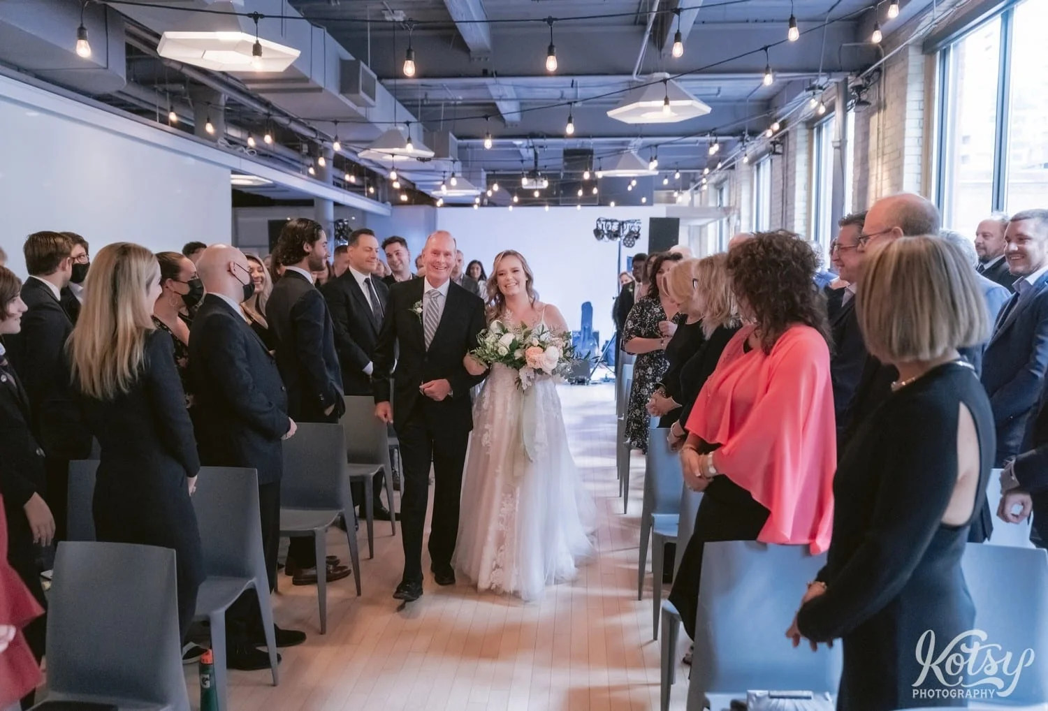 A wide shot of a bride and a white bridal gown holding a bouquet of flowers walks down the aisle with her father wearing a black suit during her Second Floor Events wedding ceremony in Toronto, Canada