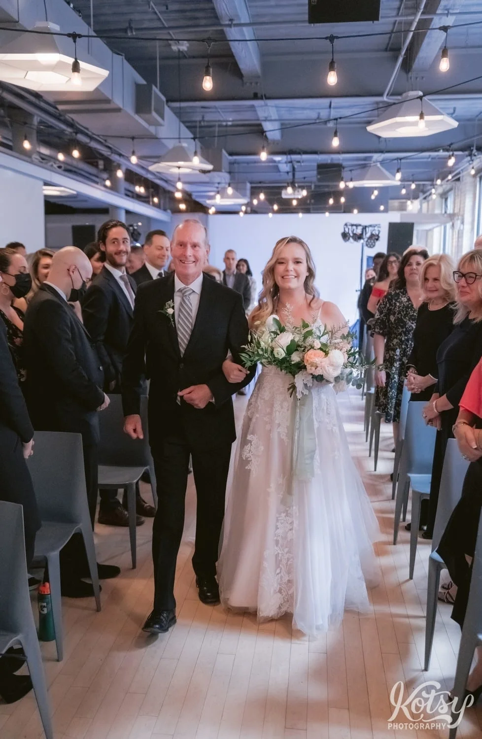 A vertical shot of a bride in a white bridal gown holding a flower bouquet walks down the aisle with her father during her Second Floor Events wedding ceremony in Toronto, Canada