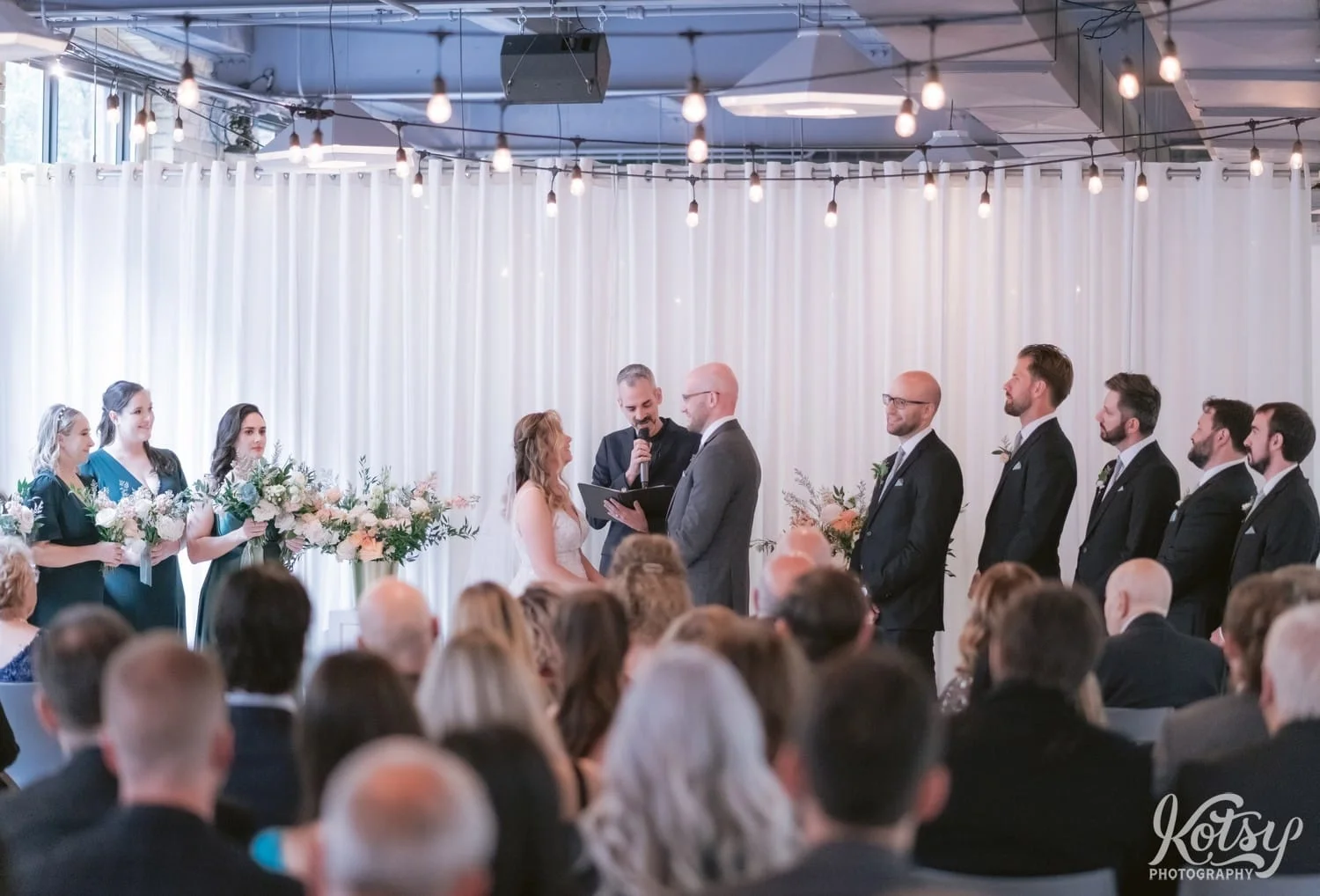 A wide shot of a bride wearing a white bridal gown and groom wearing a gray suit stand at the altar with their wedding party during their Second Floor Events wedding ceremony in Toronto, Canada