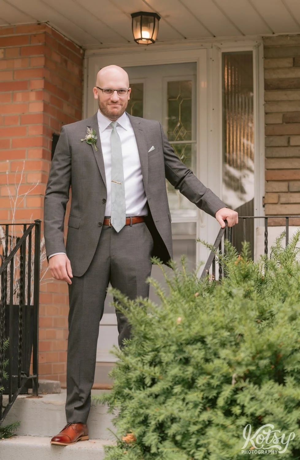 A groom wearing a gray suit and green tie stands on his front steps with his hand on the railing looking at the camera with a smile