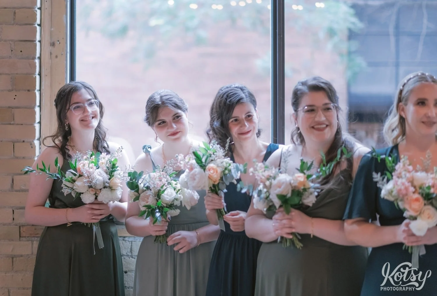A row of bridesmaids and green dresses holding flower bouquets watch with smiles during a Second Floor Events wedding ceremony in Toronto, Canada