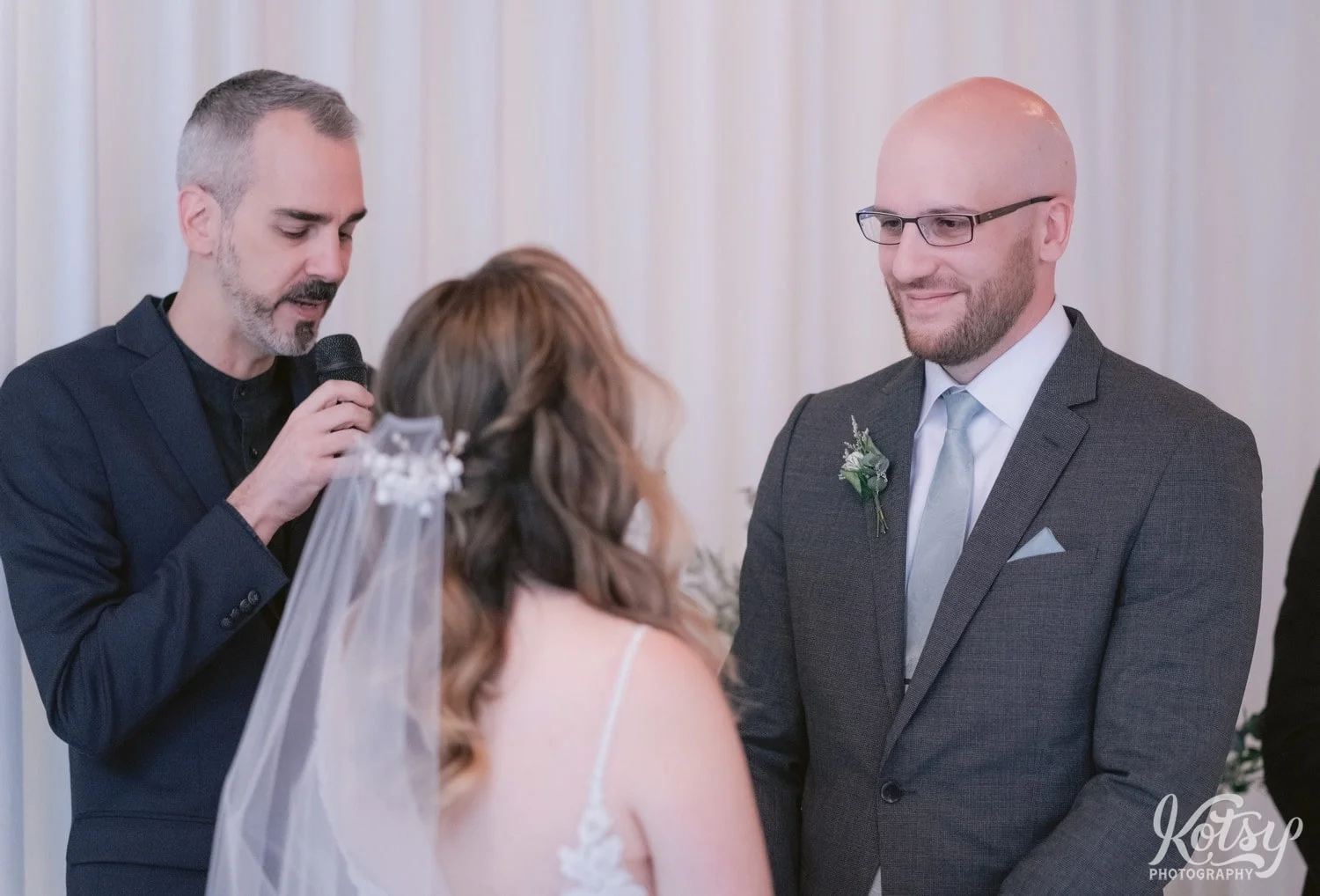 A groom in a gray suit and green tie smiles while looking at his bride as an officiant speaks into a microphone at the altar during there Second Floor Events wedding ceremony in Toronto, Canada