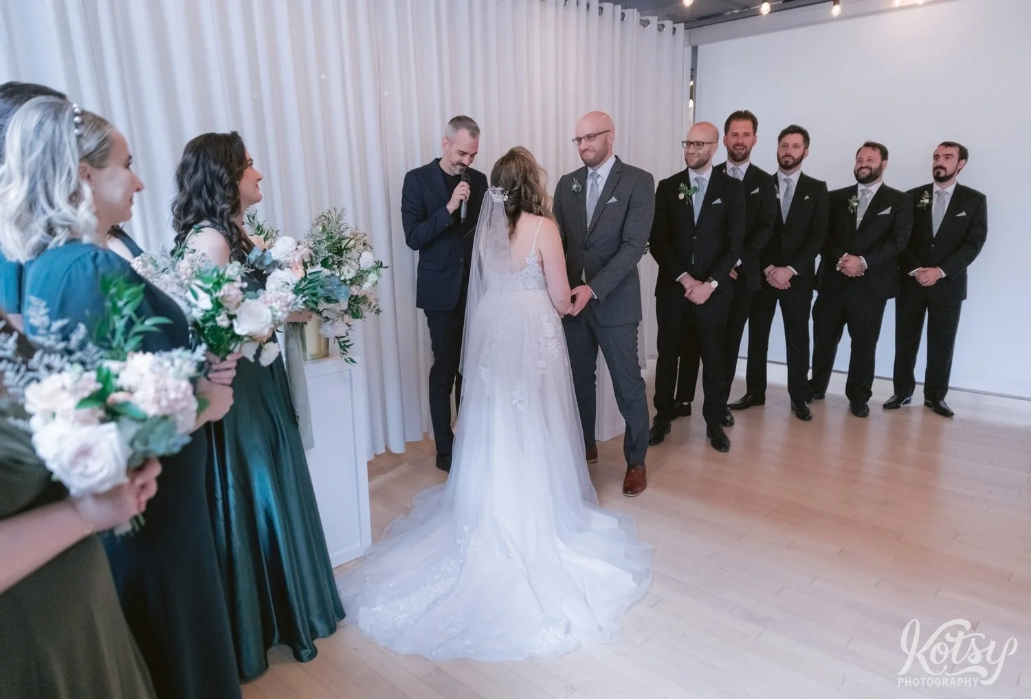 A wide shot of a groom in a gray suit and green tie looking at his bride in a white bridal gown with groomsmen behind him during his Second Floor Events wedding ceremony in Toronto, Canada