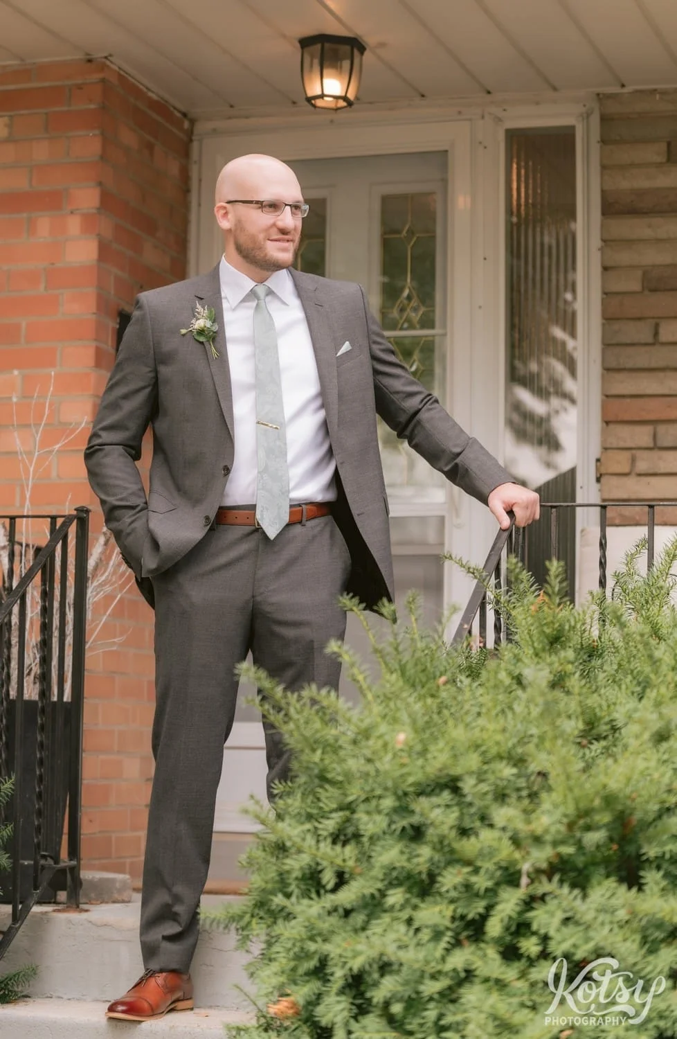 A groom standing on the front stairs to his house wearing a gray suit and green tie and his hand on the railing looking off camera