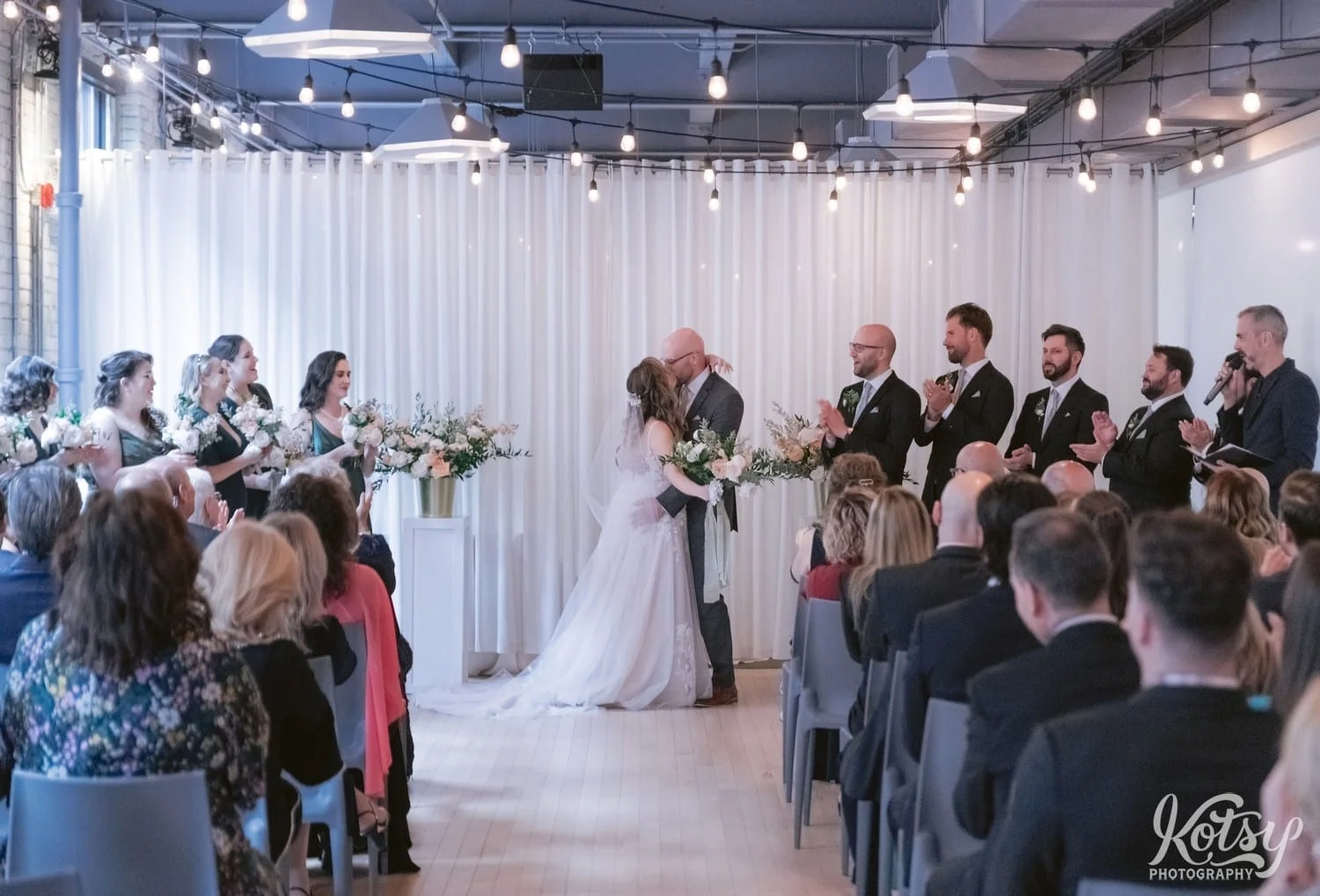 A bright and groom kiss at the altar in front of a room full of people and their wedding party during their Second Floor Events wedding ceremony in Toronto, Canada