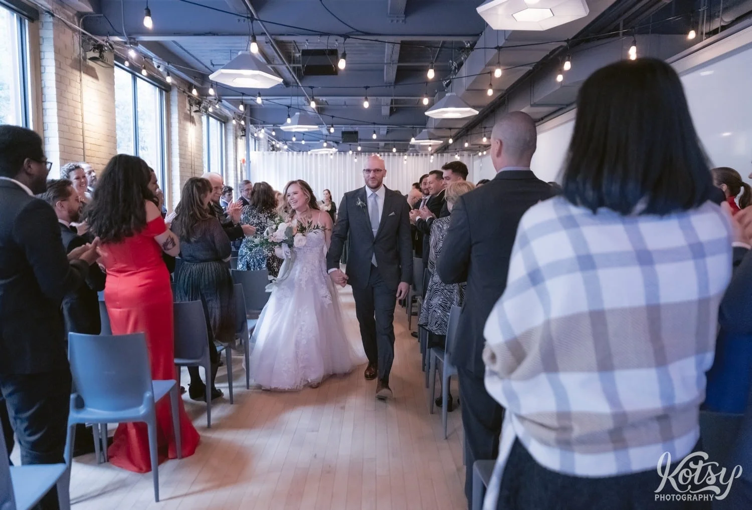 A bride in a white bridal gown walks back up the aisle holding a flower bouquet with her groom wearing a gray suit at the conclusion of their Second Floor Events wedding ceremony in Toronto, Canada