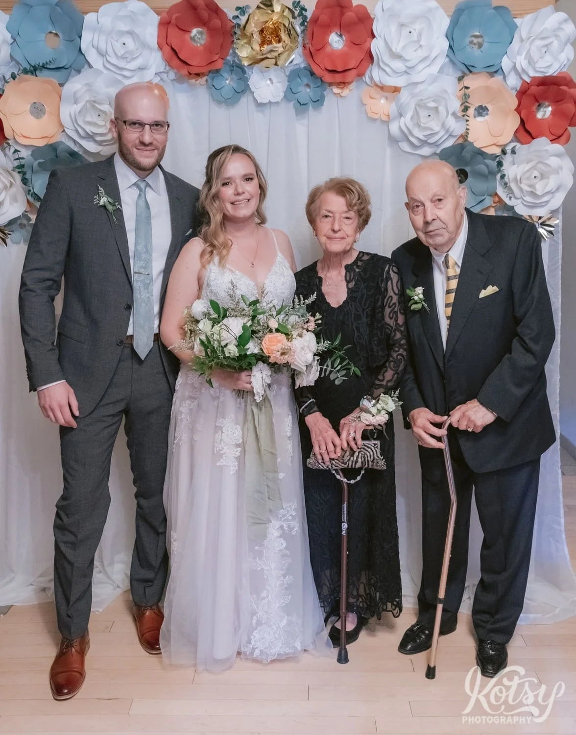 A bride wearing a white bridal gown holding a flower bouquet poses with her groom wearing a gray suit and his grandparents in front of a backdrop at their Second Floor Events wedding reception in Toronto, Canada