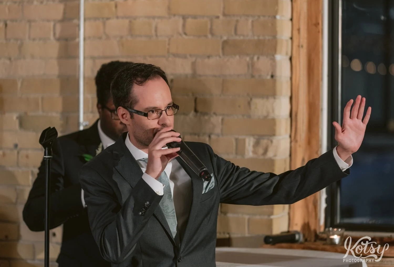 A man in a black suit and green tie speaks into a microphone with his hand in the air during a Second Floor Events wedding reception in Toronto, Canada.