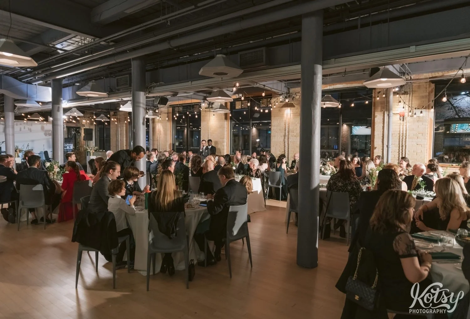 A white shot of a room full of tables and people watching a man address them during Second Floor Events wedding reception in Toronto, Canada.