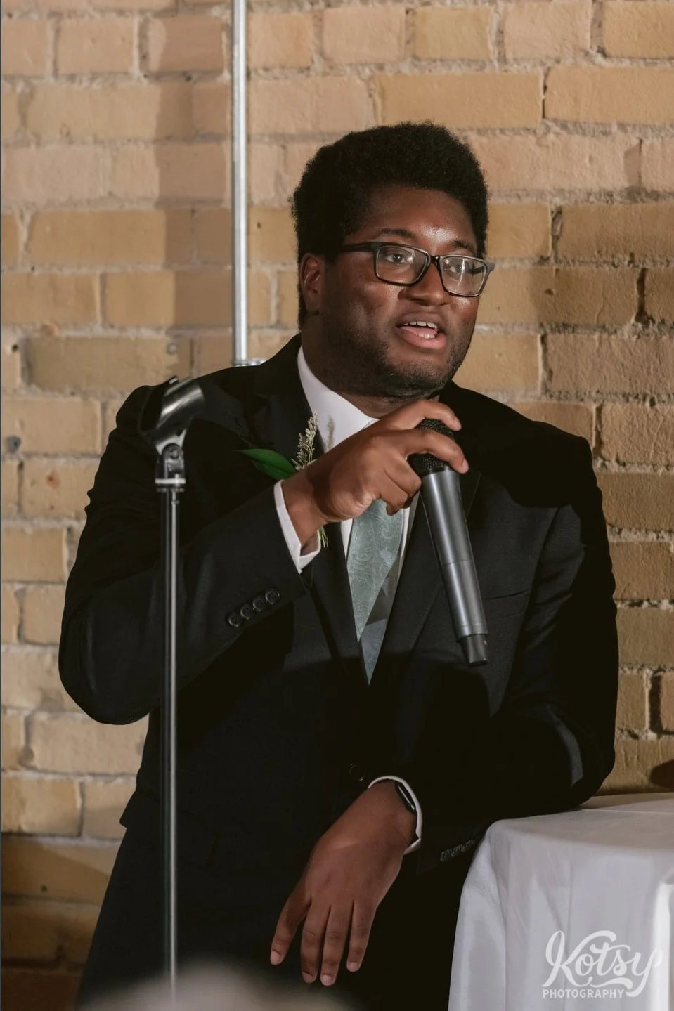 A man wearing a black suit and green tie speaks into a microphone in front of a brick wall during a Second Floor Events wedding reception in Toronto, Canada.