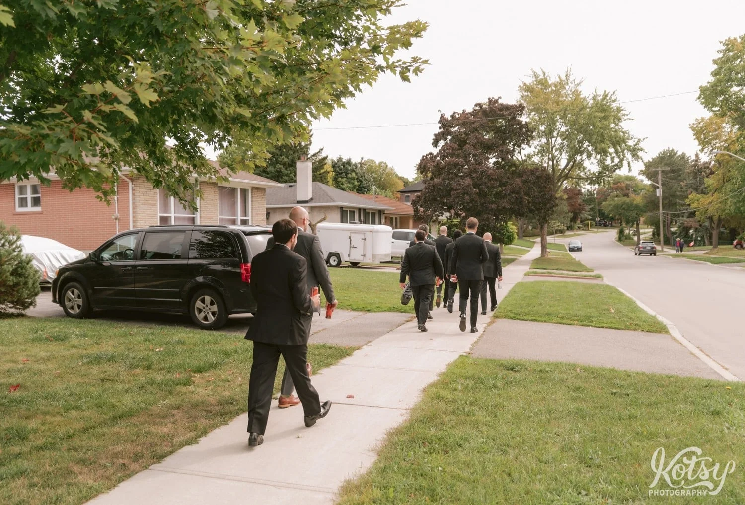 Rear view of a group of eight groomsmen and a groom walking on a sidewalk in a residential neighborhood towards a park
