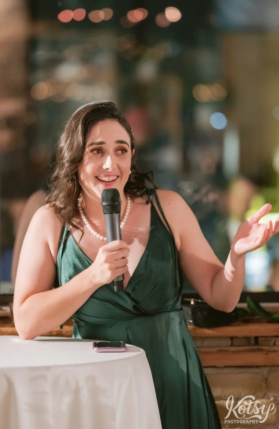 A woman in a green dress speaks into a microphone in front of a table with white cloth on it during a Second Floor Events wedding reception in Toronto, Canada.