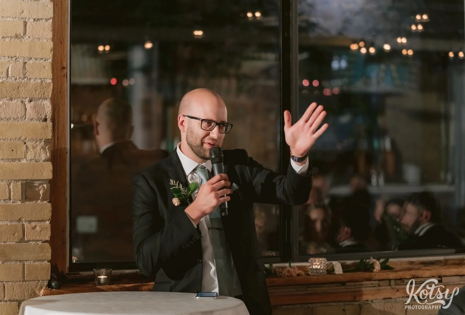 A man in a black suit and green tie and glasses speaks into a microphone with his hand raised during a wedding reception at second floor events in Toronto Canada