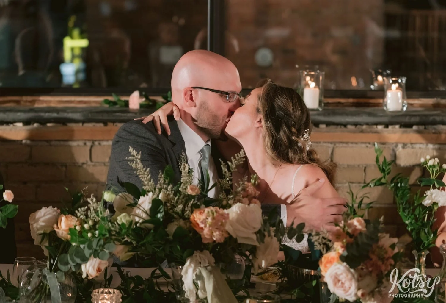 A bride and groom enjoy a kiss while sat at a dinner table behind a large setup of flower bouquets during their Second Floor Events wedding reception in Toronto, Canada.