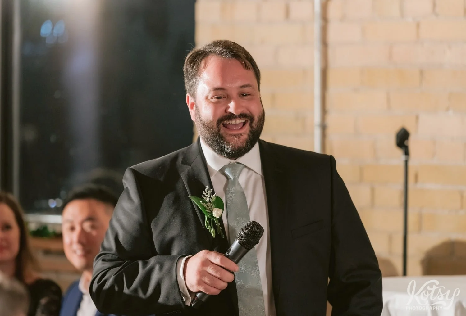 A man in a black suit and green tie enjoys a laugh while holding a microphone during a Second Floor Events wedding reception in Toronto, Canada.