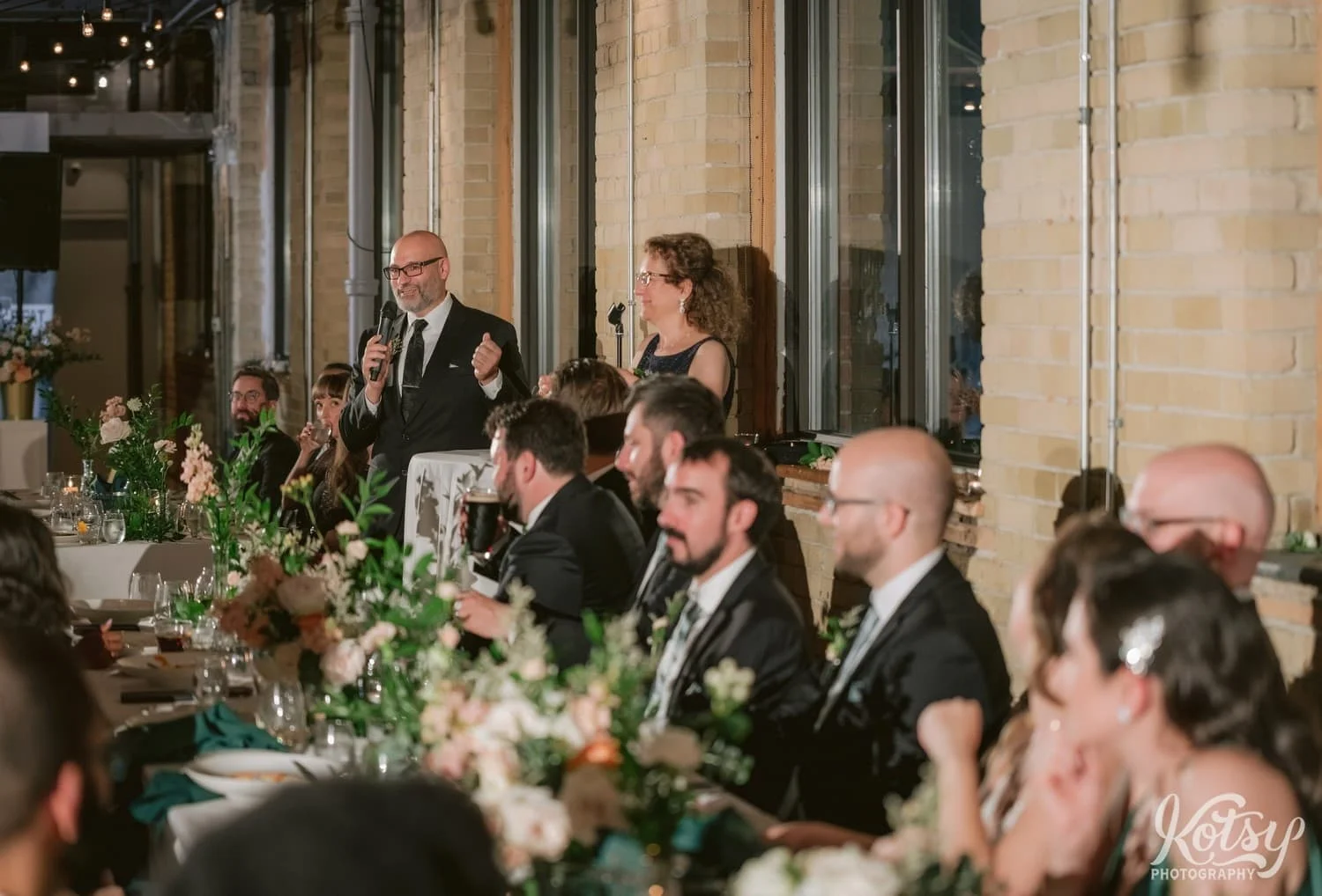 A man in a black suit and black tie speaking into a microphone is seen beyond a row of people sitting at a long table during a Second Floor Events wedding reception in Toronto, Canada.