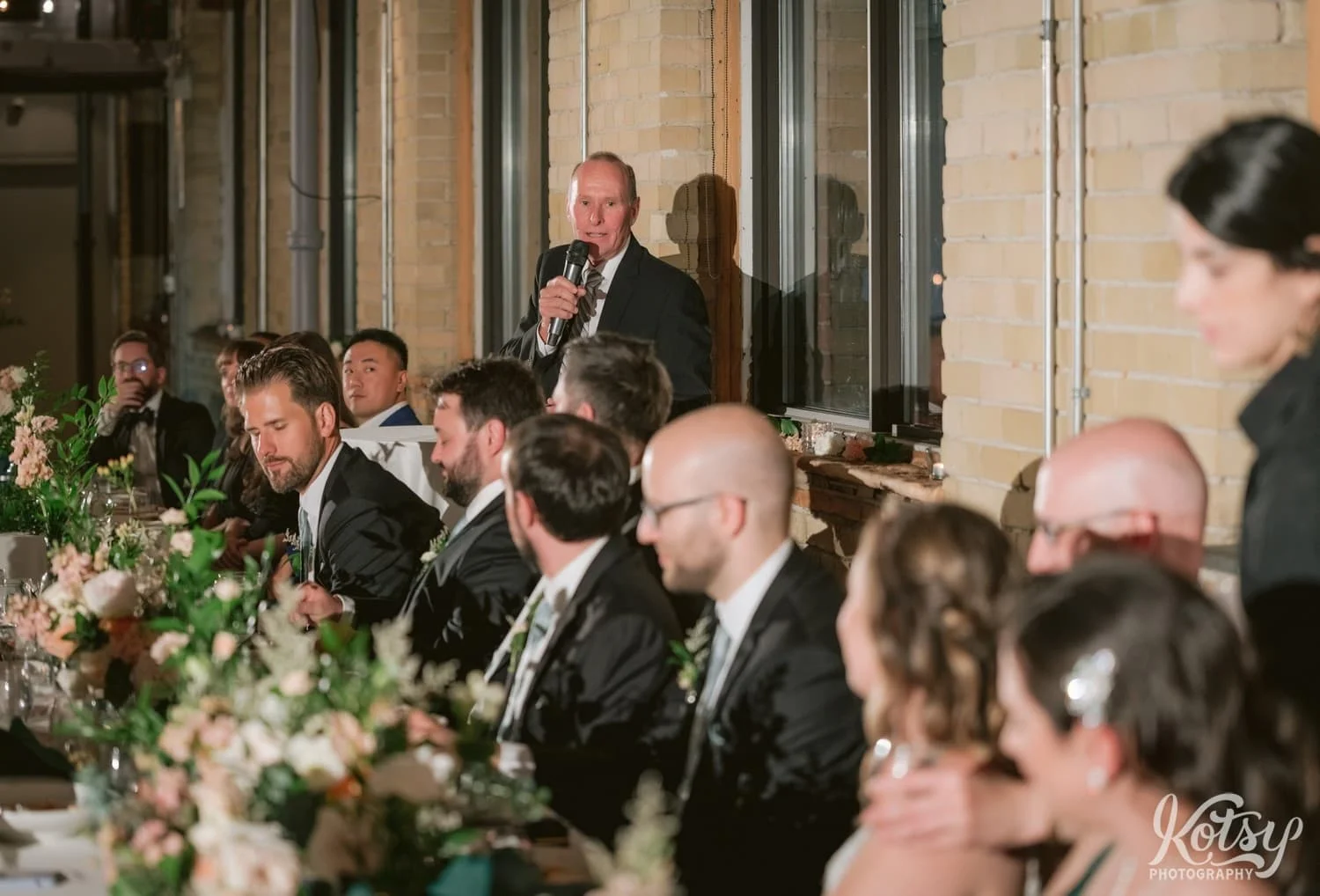 A man in a black suit holding a mic makes a speech to a room full of people during a Second Floor Events wedding reception in Toronto, Canada.