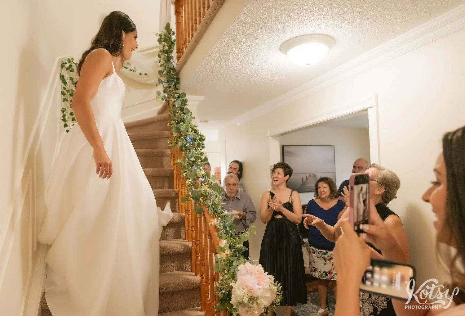 A bride is all smiles as she comes down a set of stairs inside a house with her family at the bottom