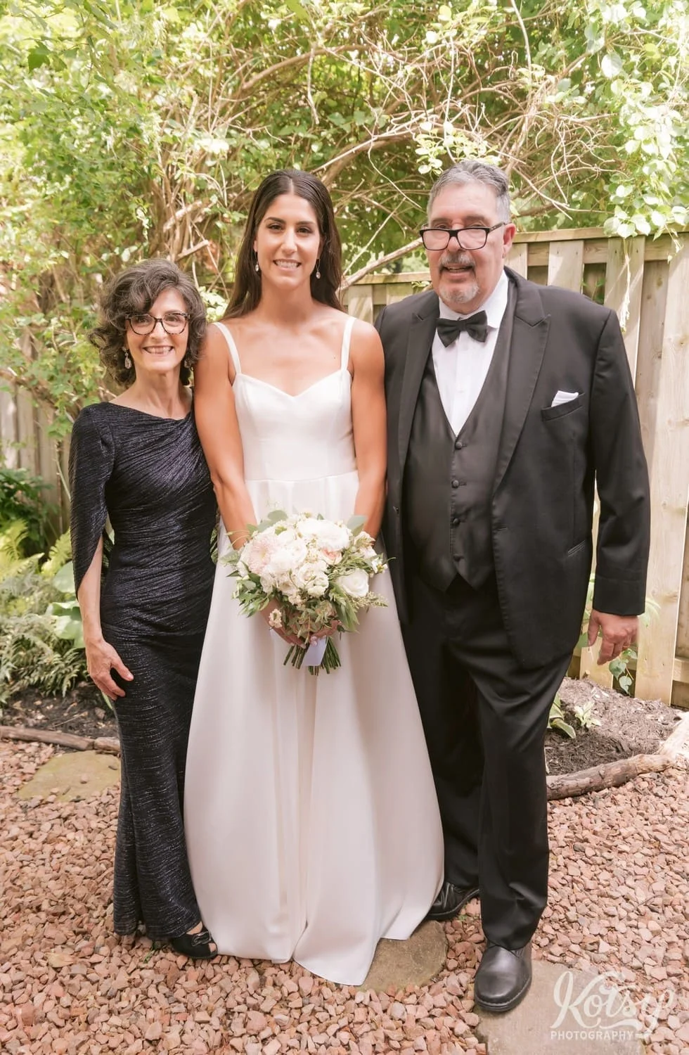 A bride wearing a white wedding down and holding a bouquet of flowers poses for a photo with her parents in a backyard