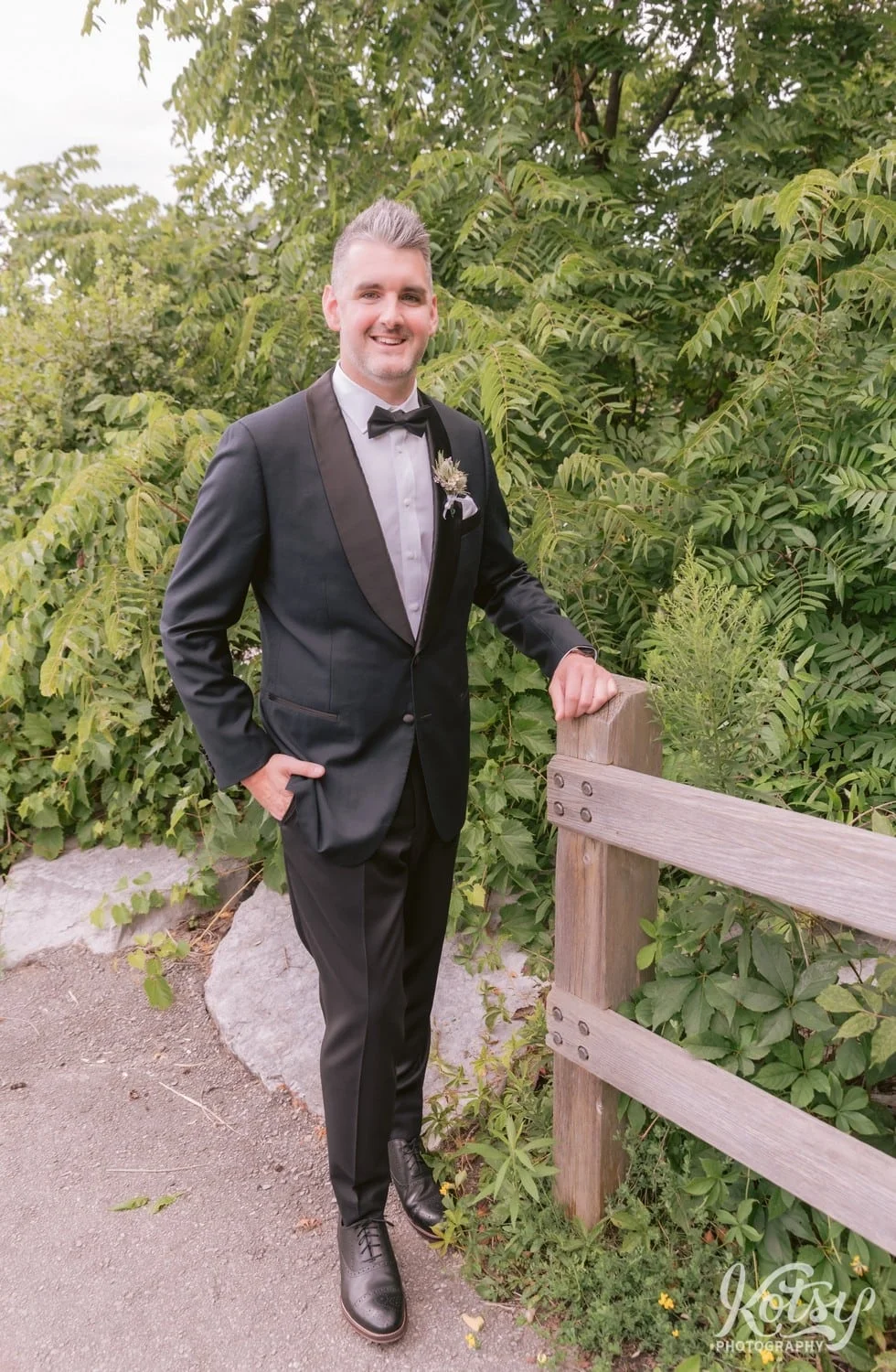 A man in a back tuxedo poses for a photo with one hand in his pocket and the other on a wooden fence post